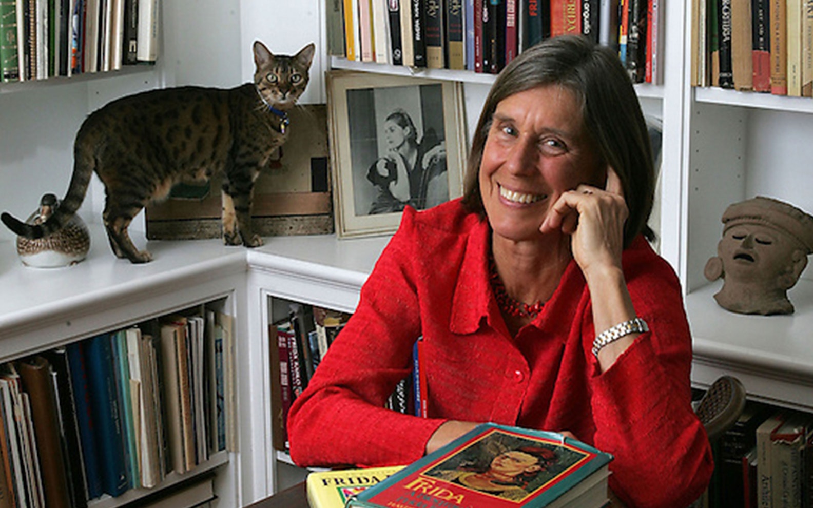 Hayden-Herrera An adult with light skin, wearing a red shirt and silver watch, has medium legth brown hair and smiles at the viewer with a hand to their face. A cat stands on a bookshelf behind.