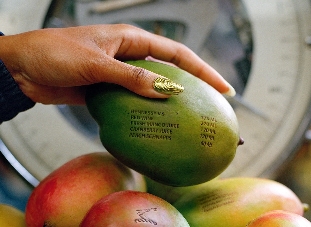 A hand with gold nails holds a mango with cocktail ingredients printed on its skin, above more mangoes.