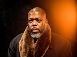Portrait of a man with dark skin in a black wool coat with brown fur collar. There is an orange flare over the upper right corner.