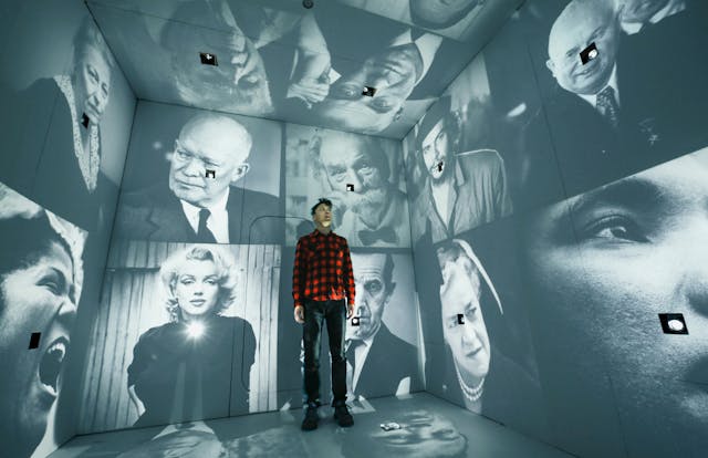 Man standing inside cube with black and white projections of portraits on every surface