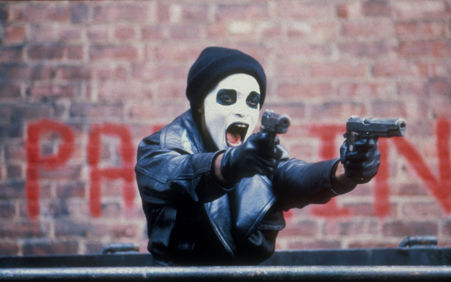 A man in a leather coat, stocking cap, and white and black face make up yells as he point guns in both hands at something off screen.