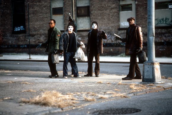 A group of men, two with white face make up on, stand at a street corner holding guns and bags of money.