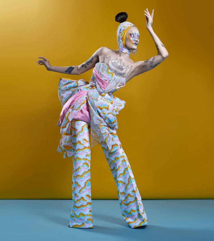 A person wearing colorful geometric makeup and garment with cloud like pattern poses against at yellow backdrop