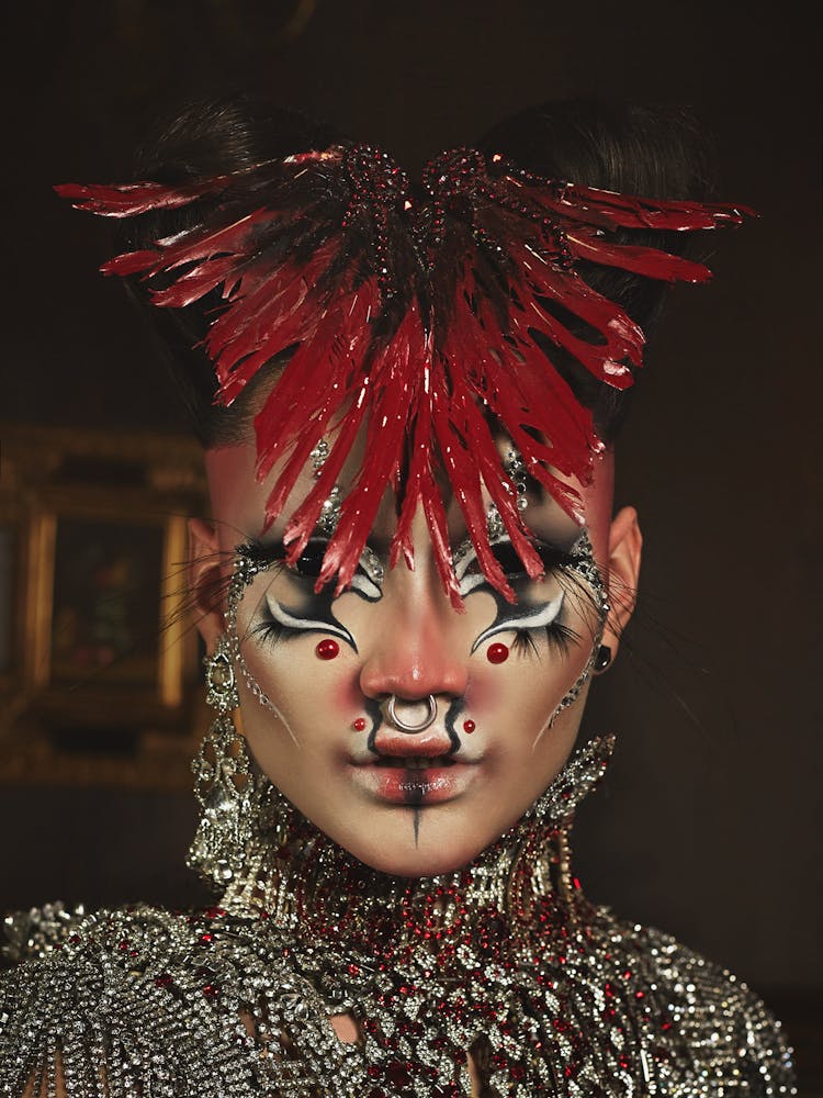 A person wearing geometric alien-like make-up and a feather headpiece looks at the viewer.