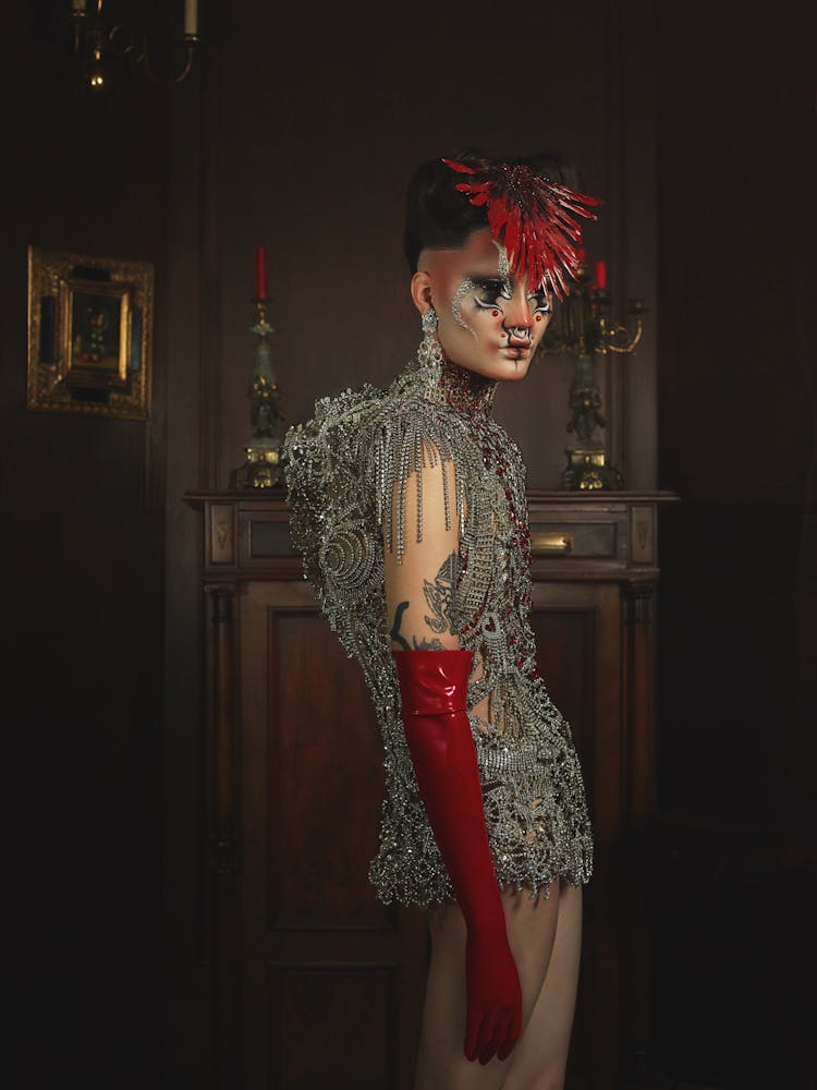 A person wearing geometric alien-like make-up, feather headpiece, and metal beaded dress stands in a dark wooden room and turns to look at the viewer.