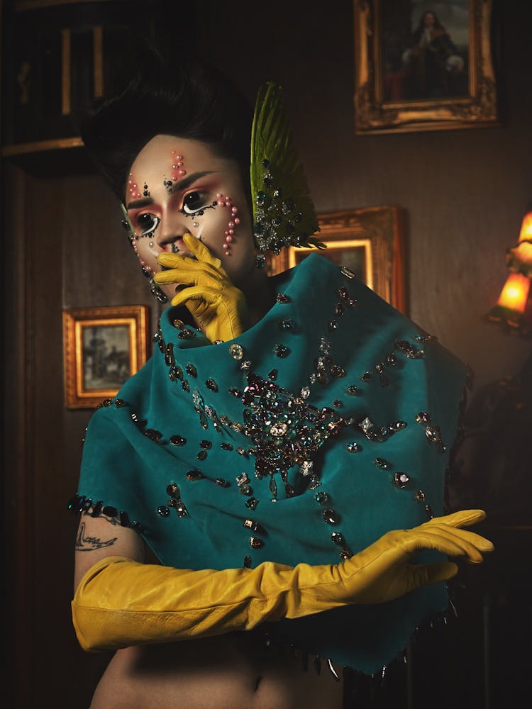 A person wearing geometric alien-like make-up, feather fins on their ears, yellow arm-lenth gloves, and a beaded shaw pulls one around through the neck hole of the shawl and looks away into the distance.