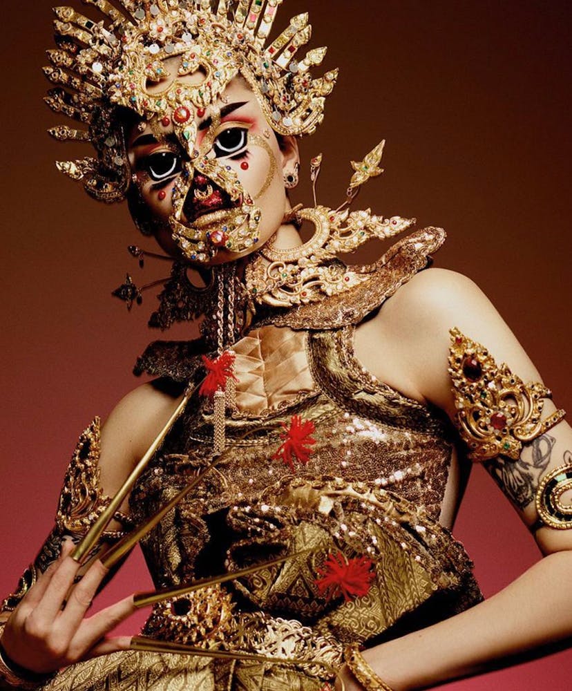 A person wearing geometric alien-like make-up and a very elaborate golden costume looks at the viewer.