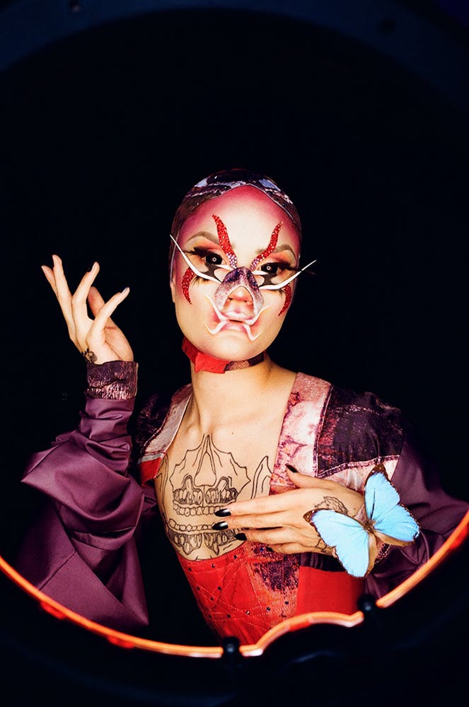 A person wearing geometric alien-like make-up holds up a hand while the other has a butterfly pirtched on it and looks at the viewer.