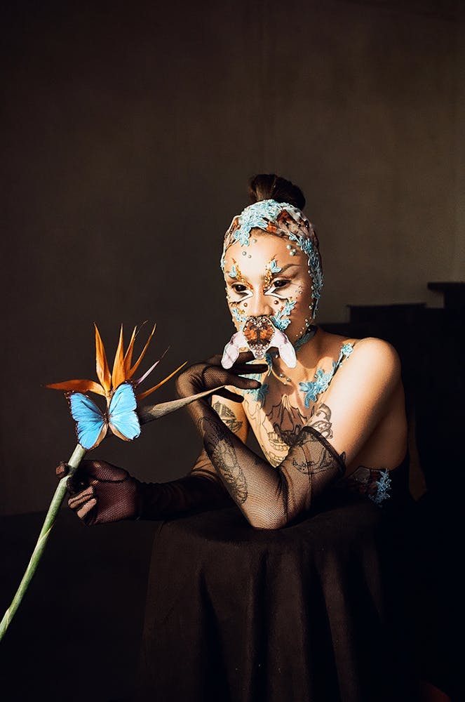 A person wearing geometric alien-like make-up holds a bird of paradise flower with a blue butterfly on it and looks into the distance.