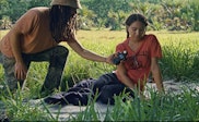 A woman with medium skin sits on a blanket in a lush green landscape while a man with medium skin leans in holding a light meter.