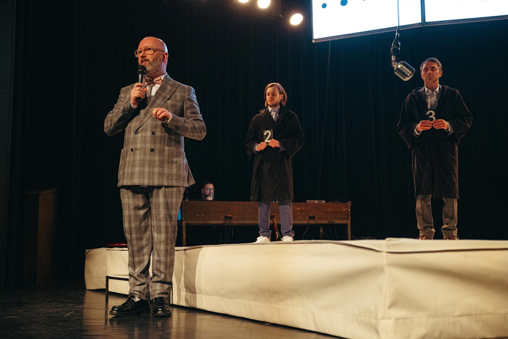 Five adults on a stage holding number signs, one in the forground facing away from the viewer holds their hand out to the others