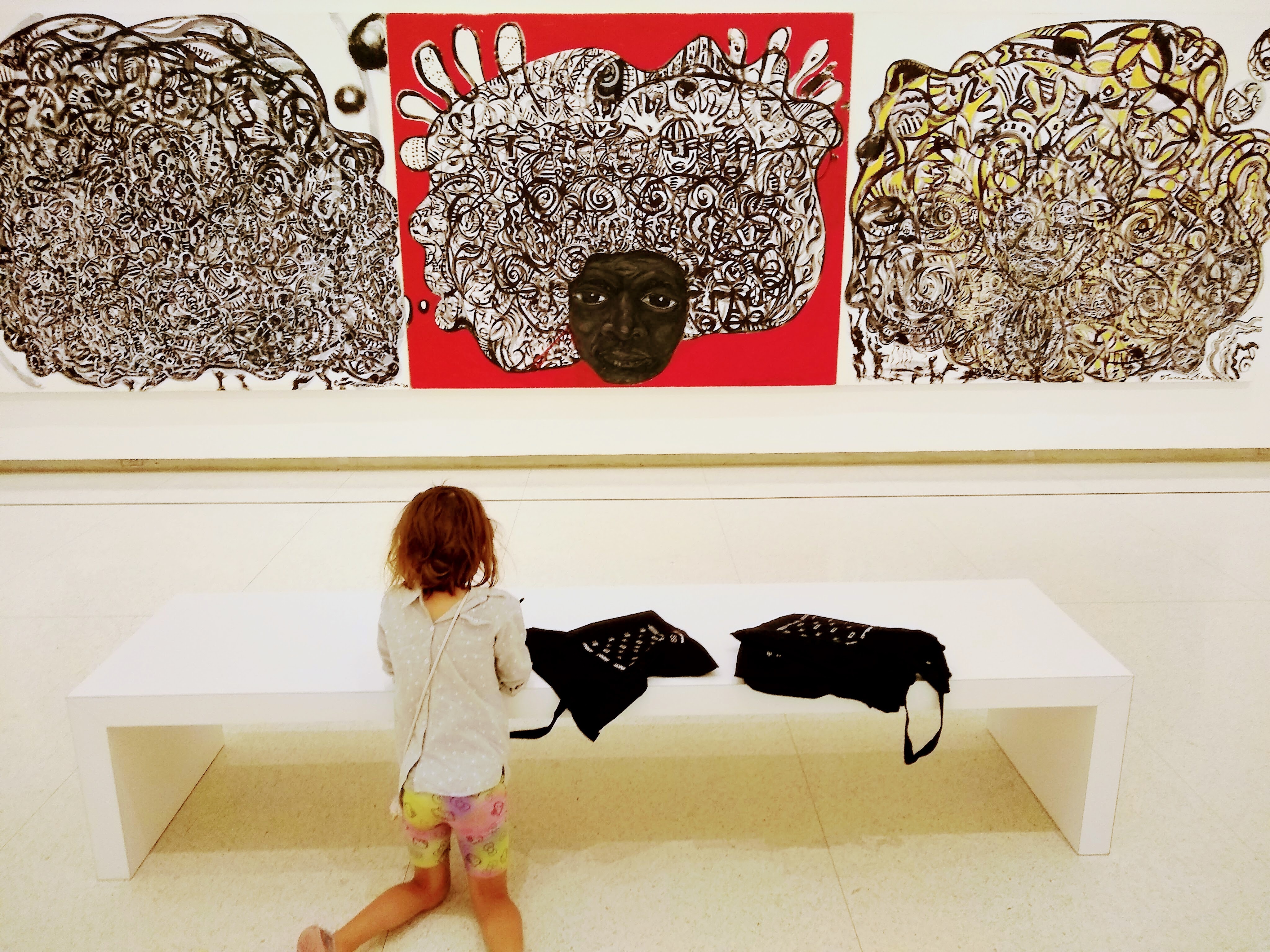A girl makes a drawing in front of a large tripich painting in a gallery.