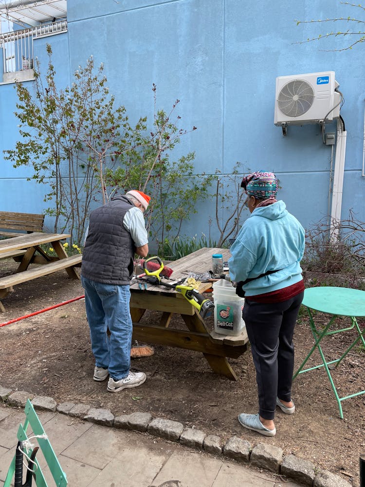 Two adults work on a weed wacker on an outdoor picnic table.