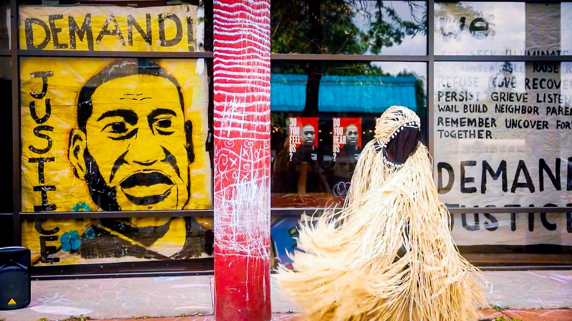 A person in a costume made of strips of straw stands outside next to a mural that reads "demand justice" and has a picture of George Flyod