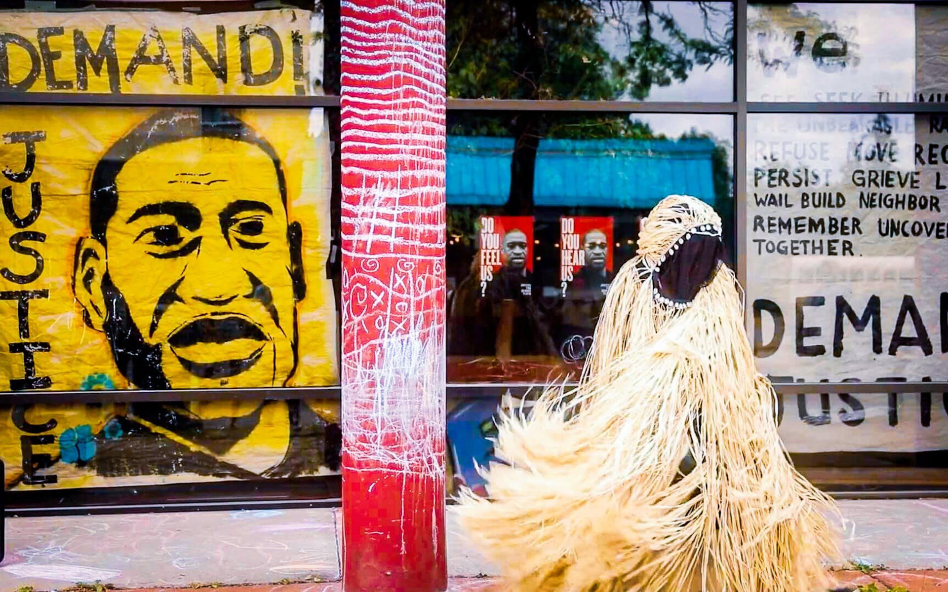 A person in a costume made of strips of straw stands outside next to a mural that reads "demand justice" and has a picture of George Flyod