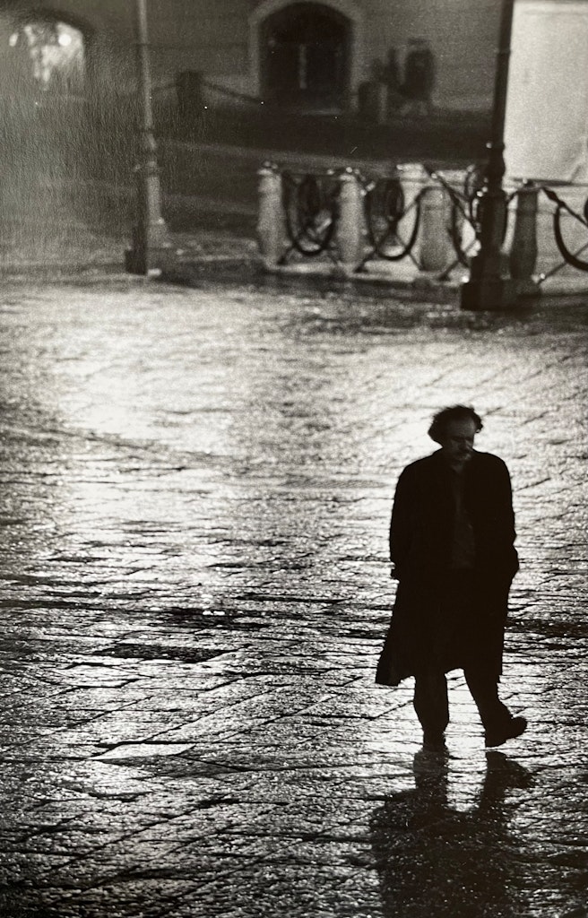 Black and white photo of a man in a long black coat walking through an empty street in Italy.