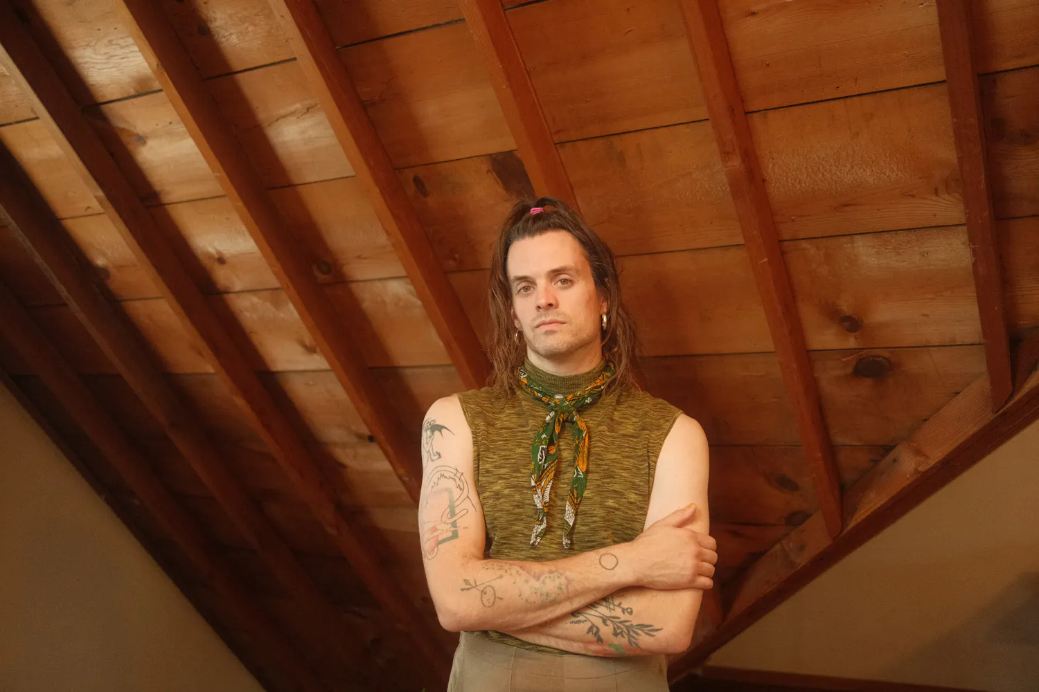 An adult with light skin tone and shoulder-length brown stands in an attic space with exposed wood beams. Their arms are crossed and tattooed.