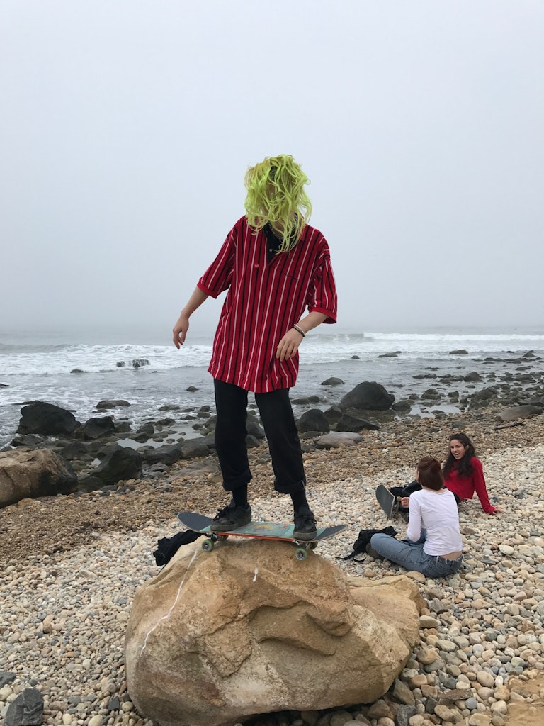 A skateboarder balances on a large rock on a beach next to the ocean near two adults siting on the ground nearby.
