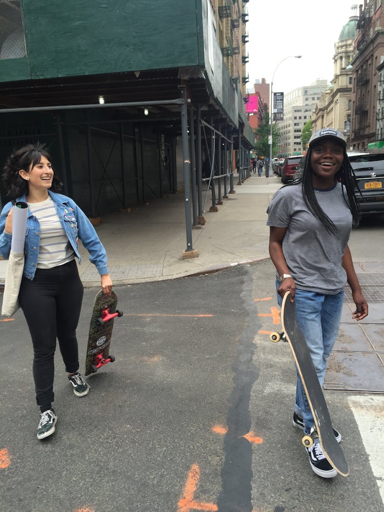 An adult with dark skin and another adult with light skin laugh while caryring skateboards acorss a city street in NYC.