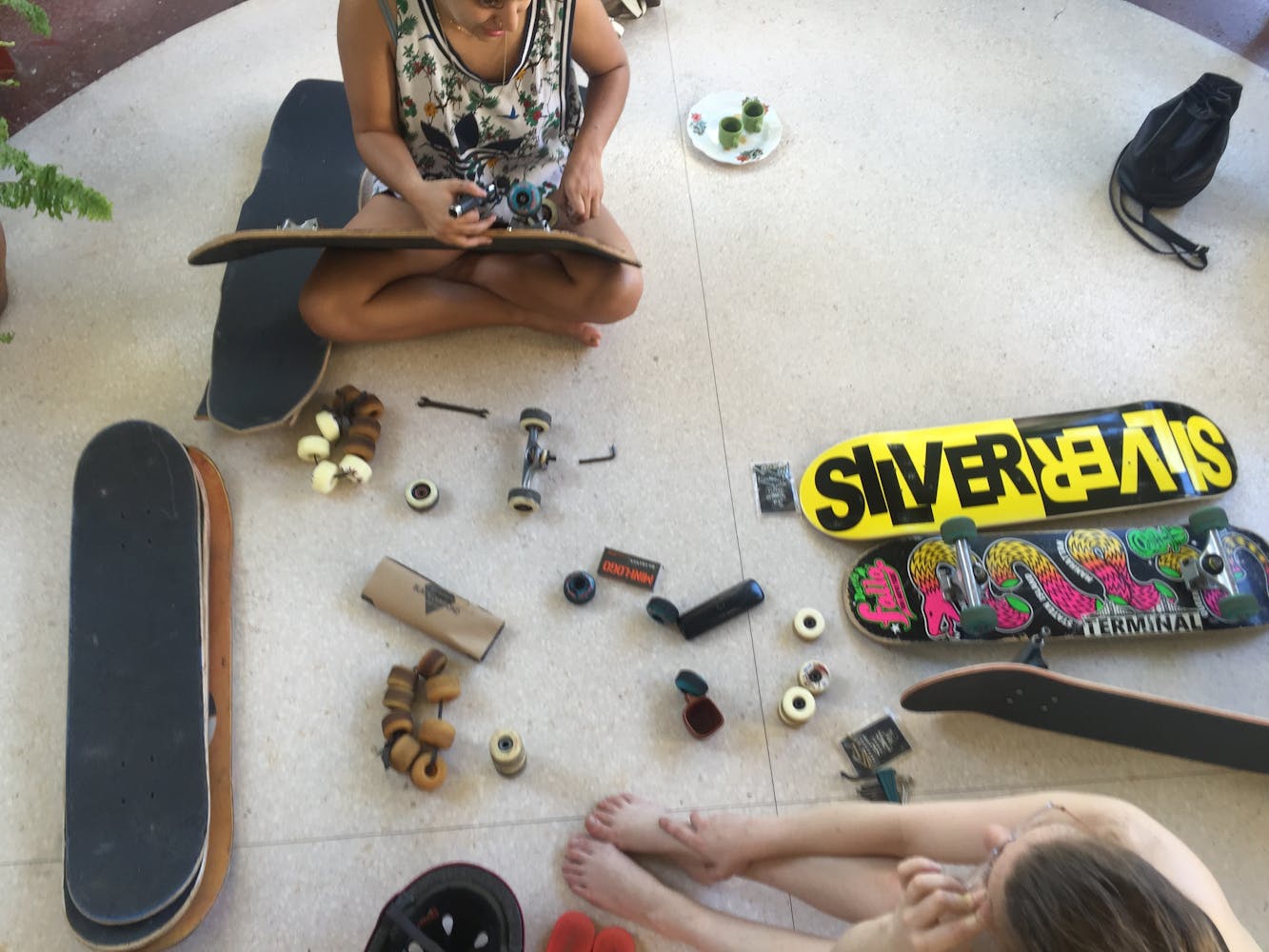 Two adults sit on a floor and place wheels on skateboards.