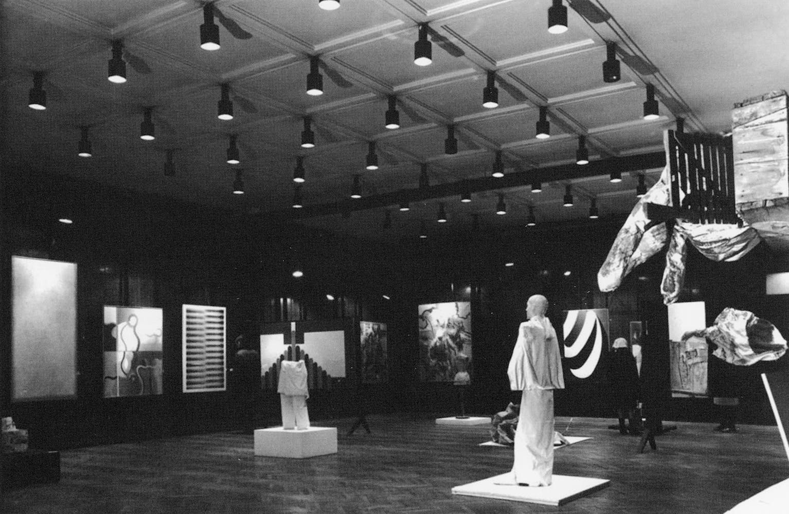 Black and white photograpgh of an art exhibition with sculptures and paintings.