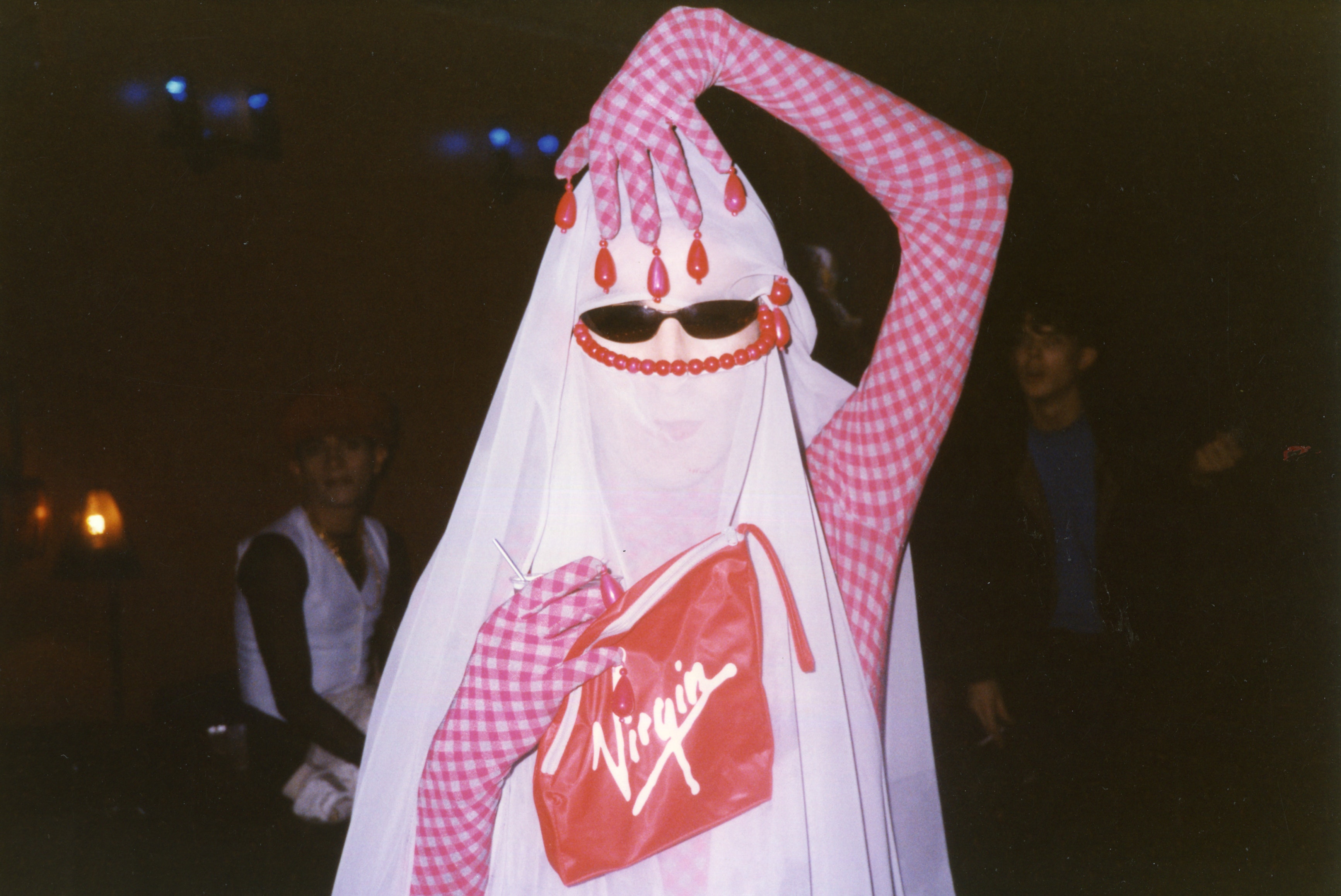An adult fully covered in a pink and white gingham print and white headscarf and sunglasses raises had above their head with beads attached to glove looks at viewer. They are holding a red bag that says 