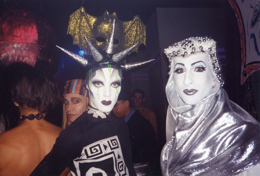 Two adults in drag make-up and silver and black costumes look at the viewer in a crowd scene.