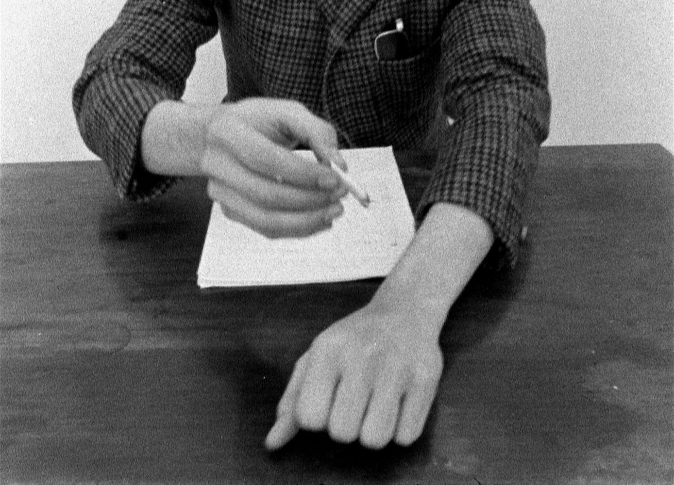 Still image looking down at someone at a table. One hand makes a fist on the table, the other holds a cigarette above the fist.