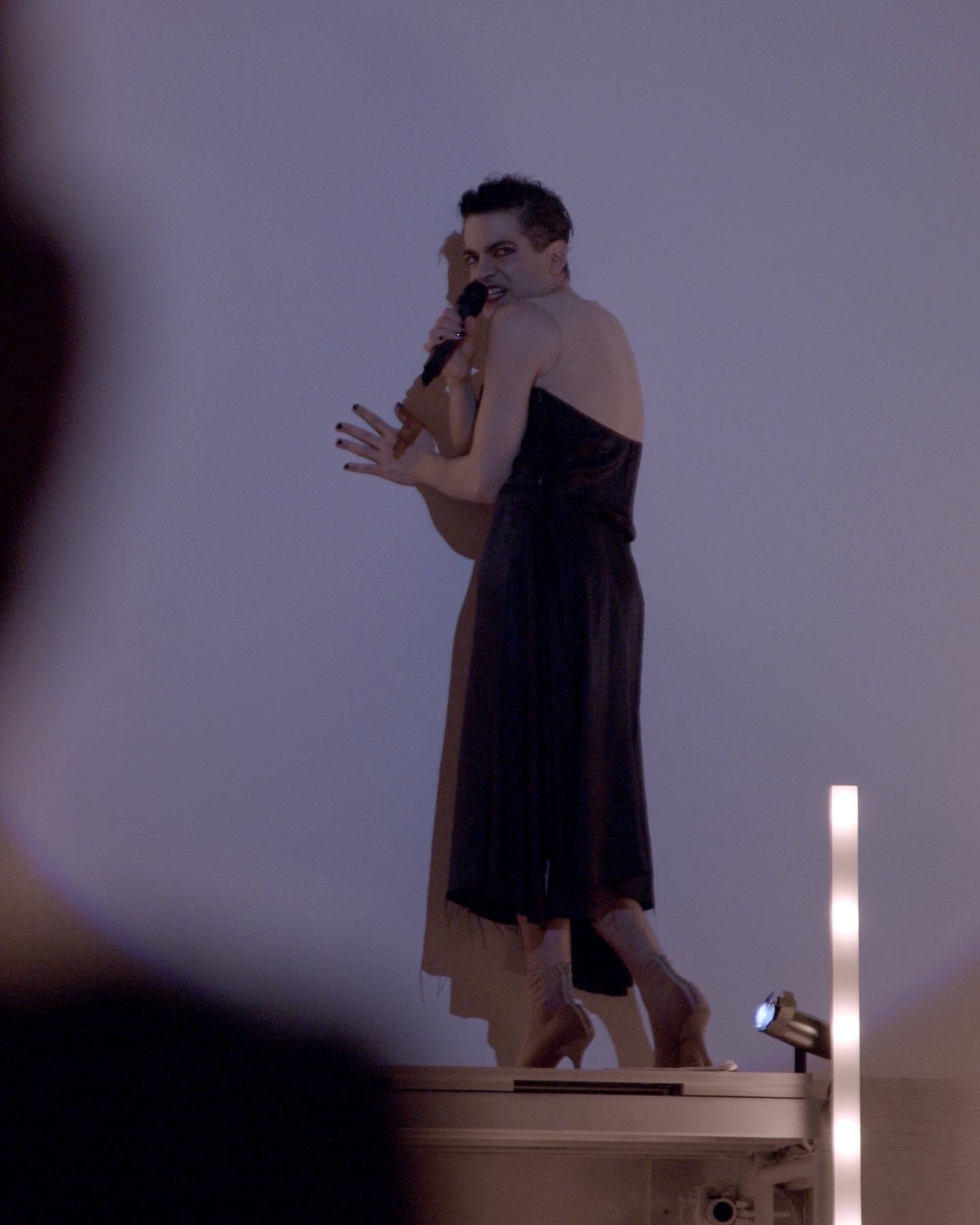 An adult in a black dress looks over their shoulder while facing a white wall and speaking into a handheld microphone.