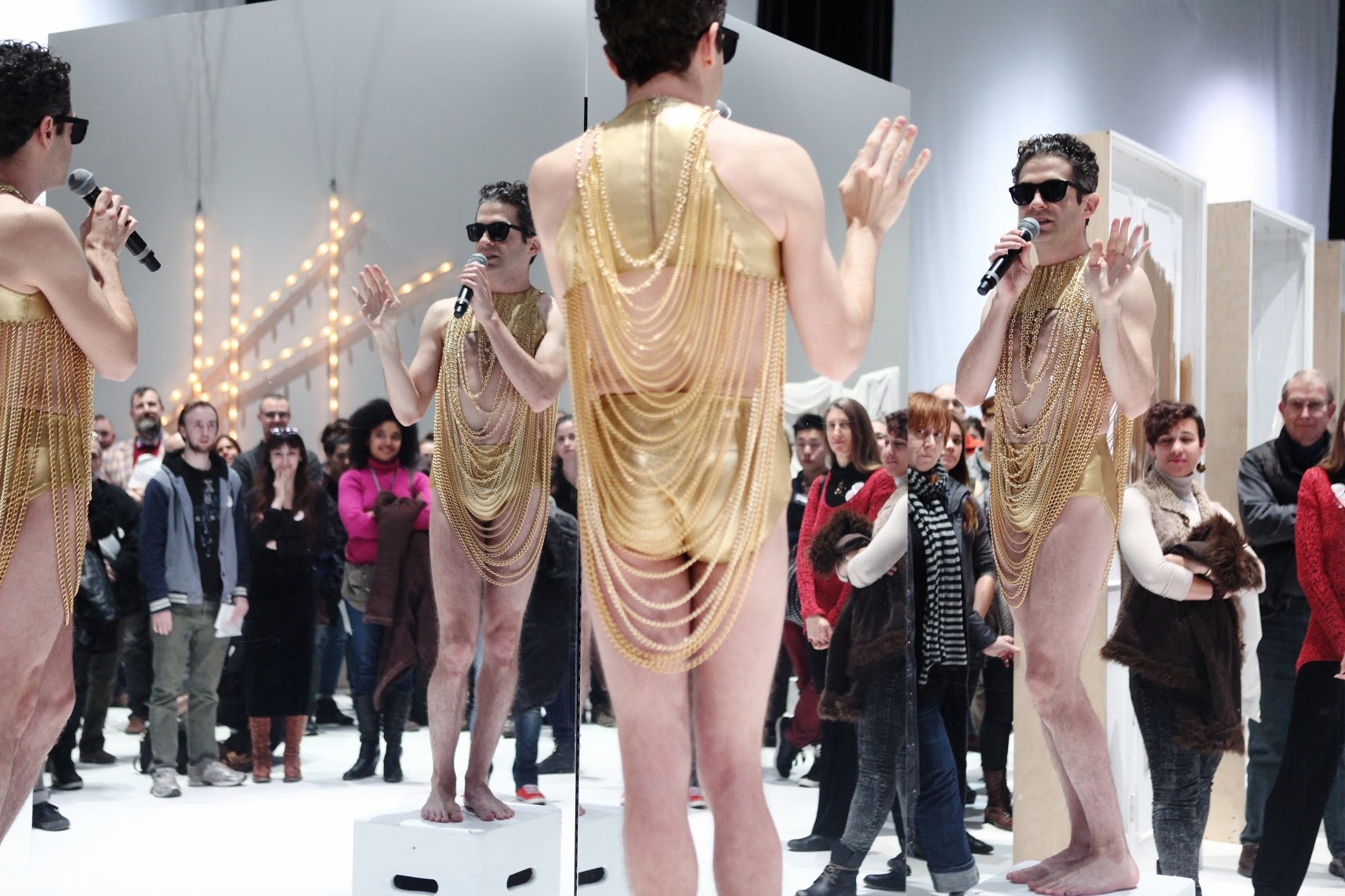And adult in glod lame underwear and tubetop with gold chains haning around their neck with black sungleasses on speaking into a microphone while looking into a corner of walls made of mirrors that reflect an audience in a gallery.