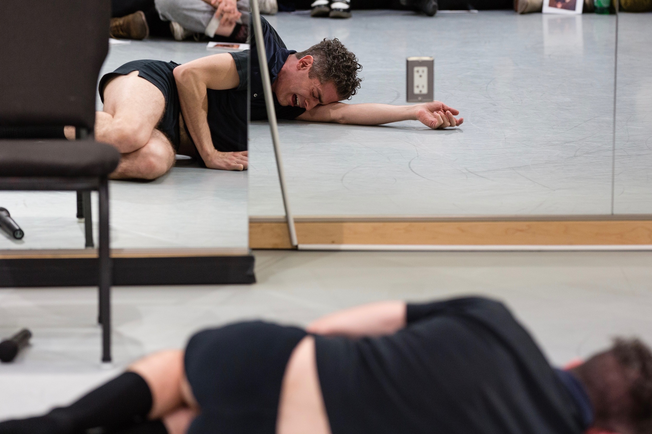An adult in dark shorts and polo shirt lies on the floor crying in front of a wall of wirros in a dance studio with an audience watching.