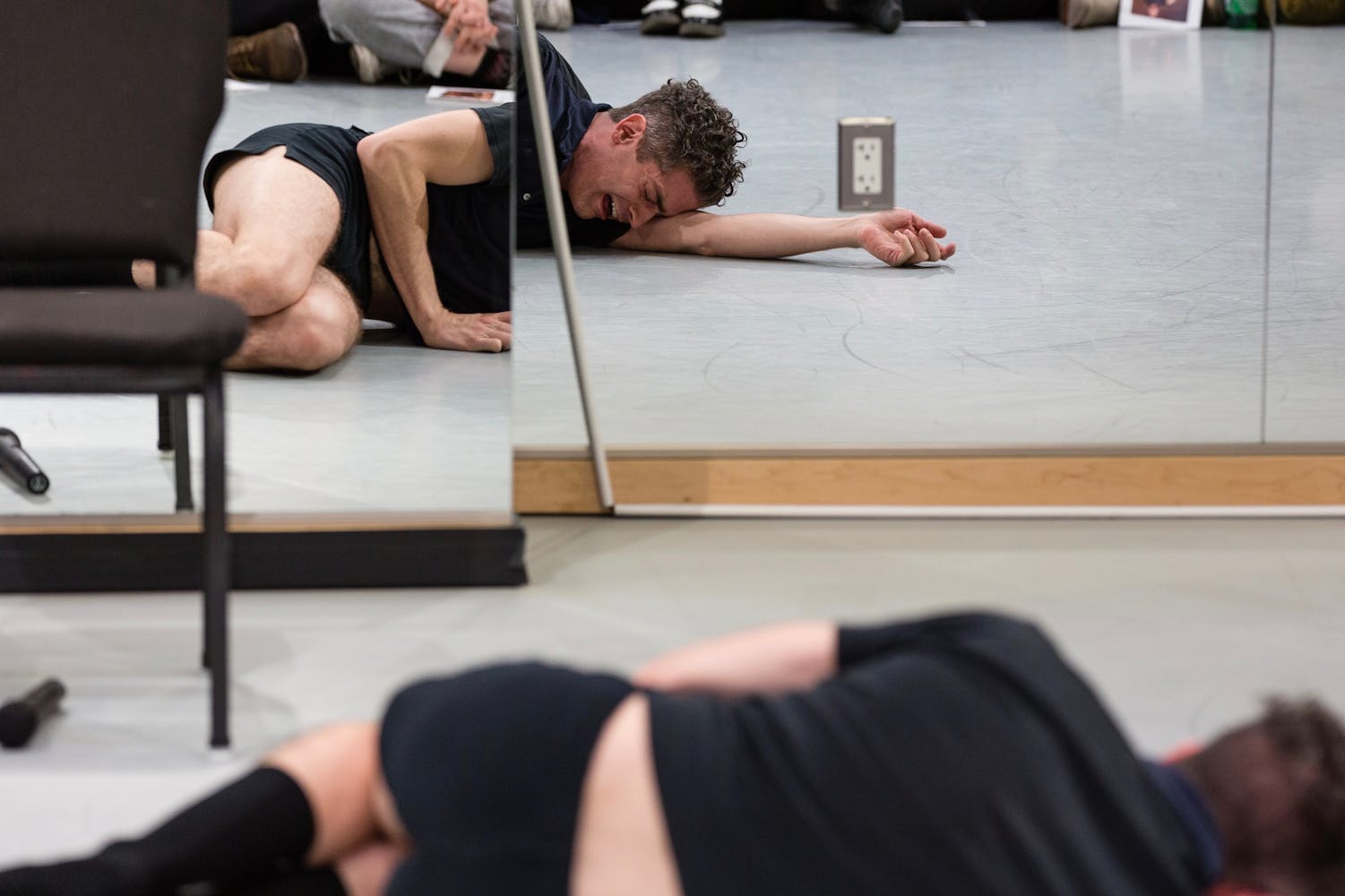 An adult in dark shorts and polo shirt lies on the floor crying in front of a wall of wirros in a dance studio with an audience watching.