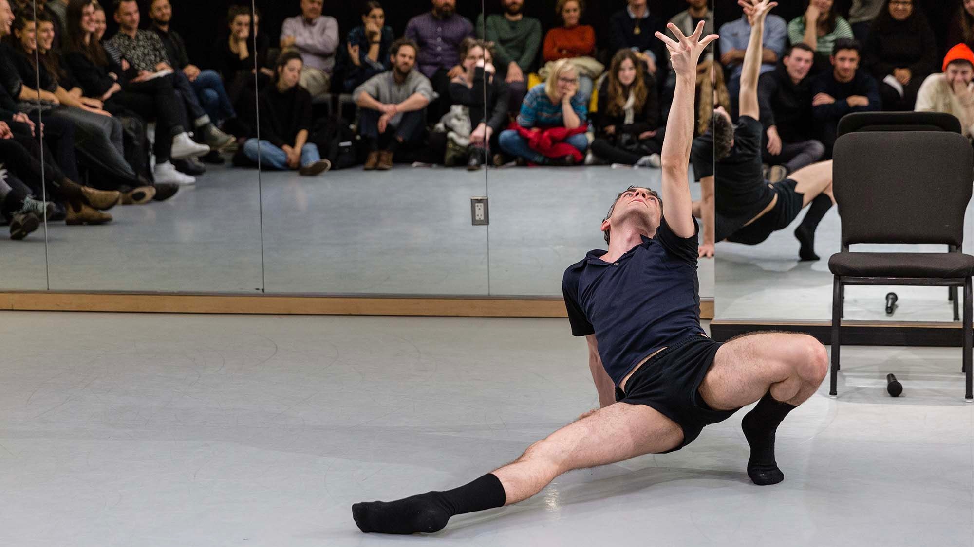 An adult in dark shorts and polo shirt poses on the floor with their left arm outstretched up into the air in front of a wall of wirros in a dance studio with an audience watching.
