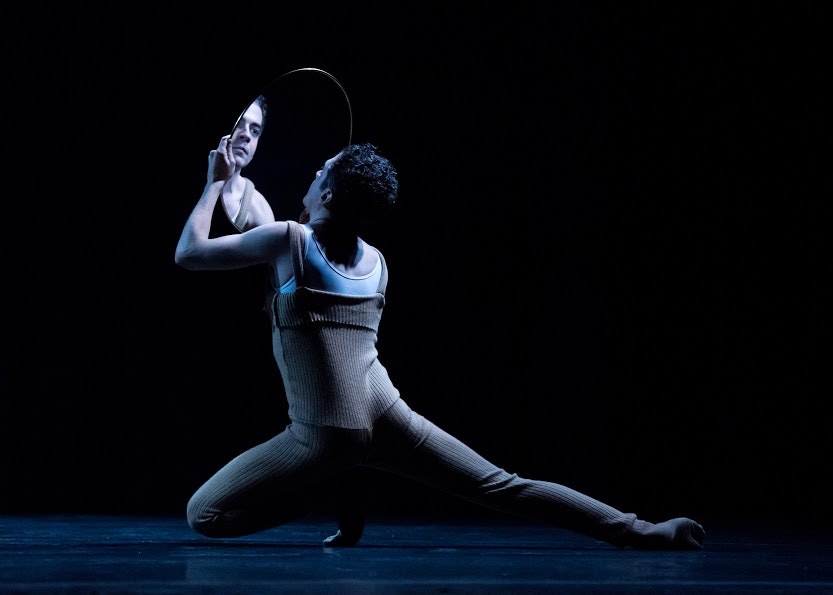 An adult in a woven body suit kneels on the ground of a black stage while looking at their reflecting in a mirror they are holding up with both hands.