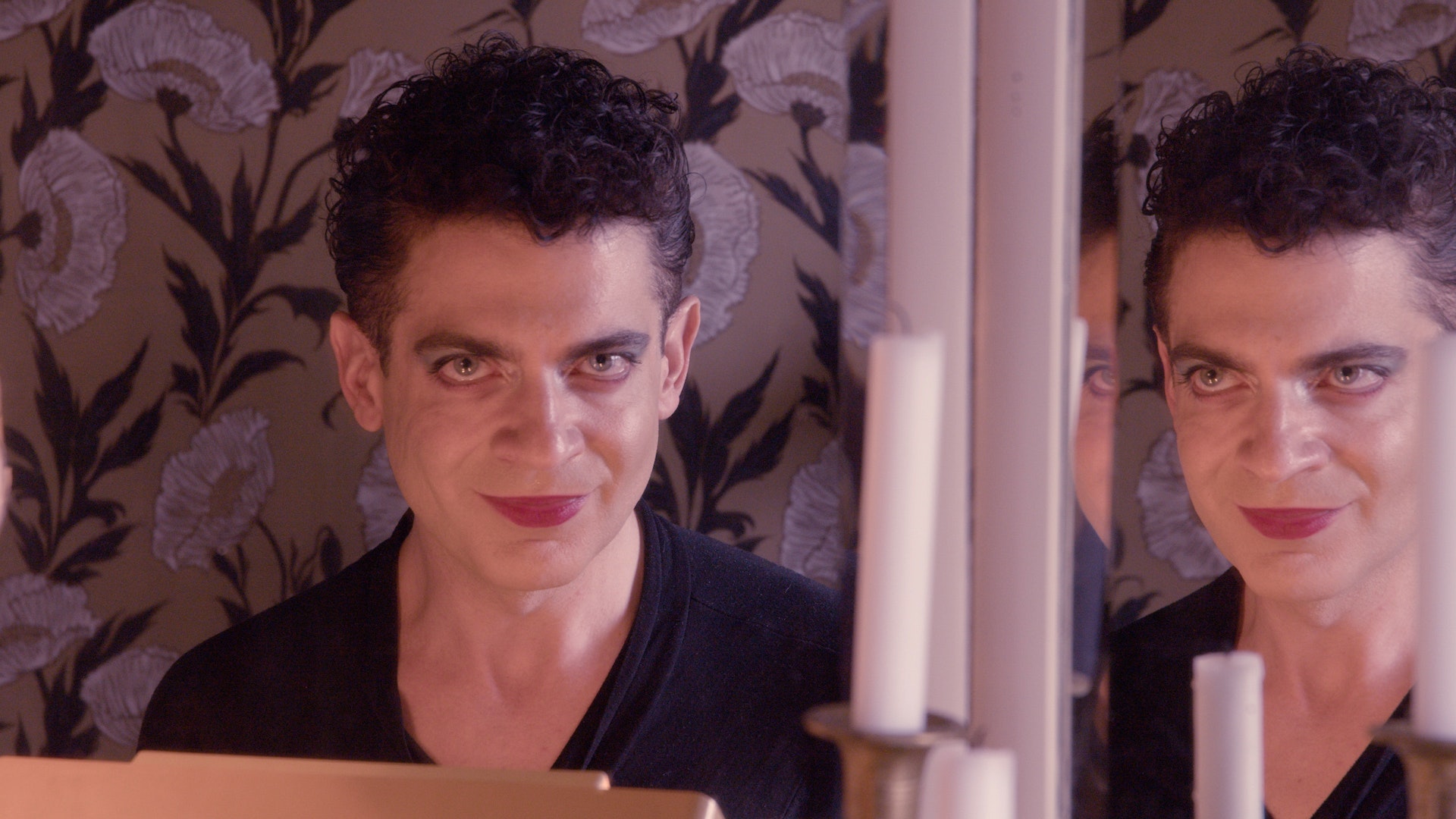 An adult wearing lipstick and eye makeup looks at the viewer with a creepy smile as a mirror next to them reflects their face.