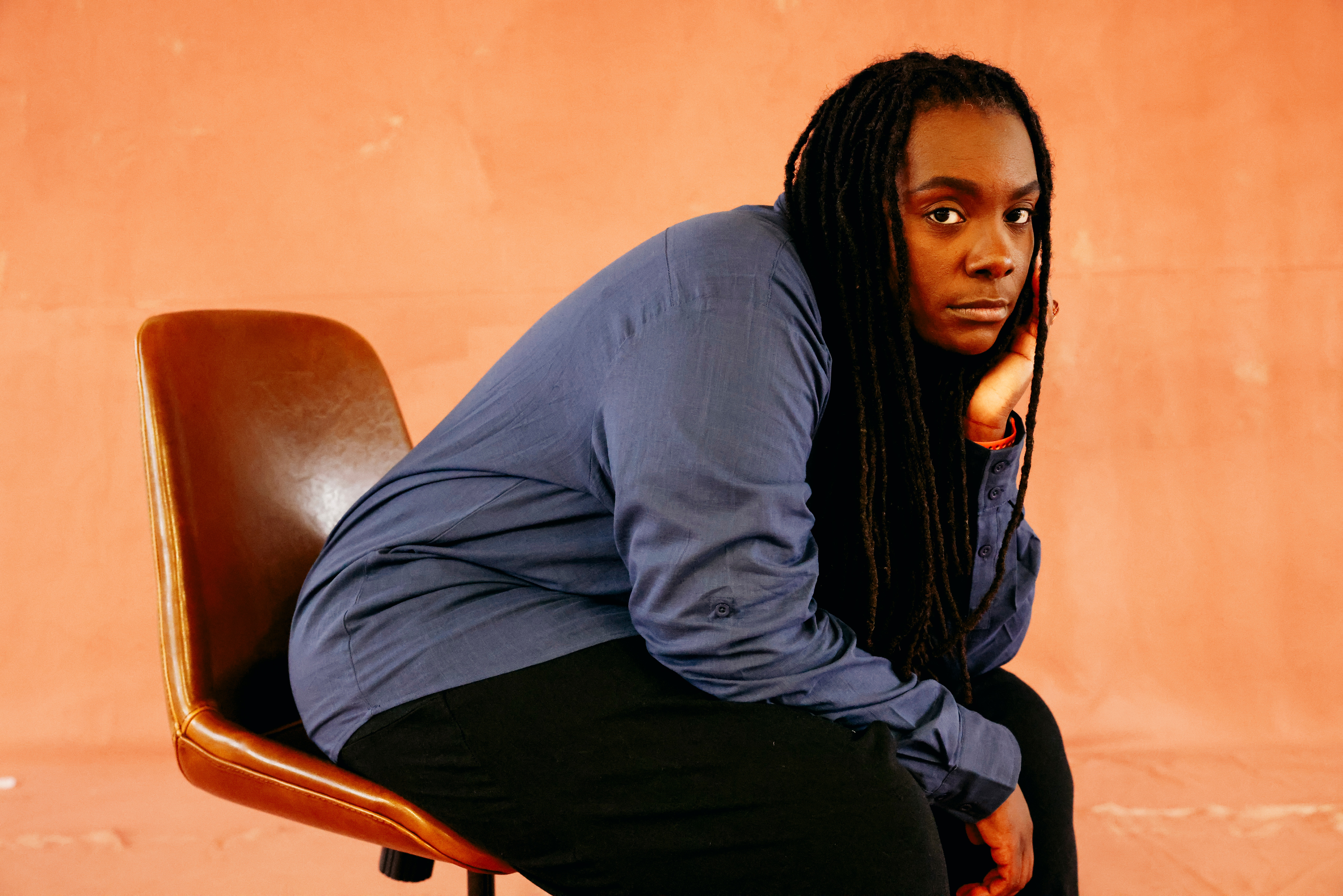 A musician with dark skin and long hair sits on a leather chair, looking forward with a neutral gaze.