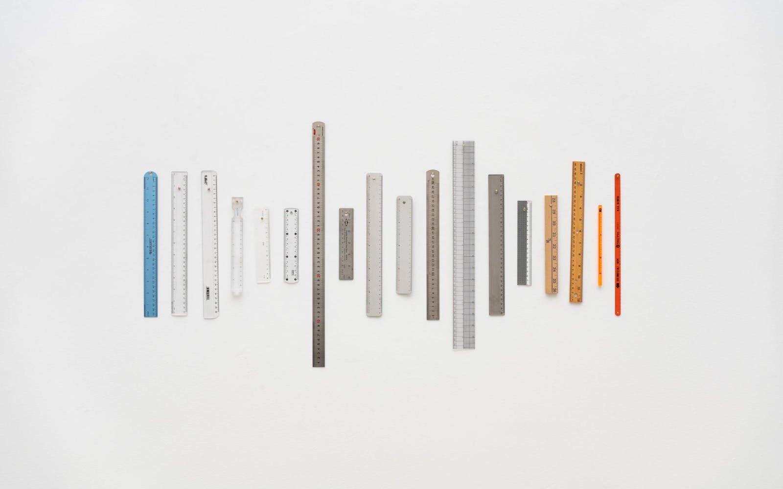 eighteen rulers of varying size ranging from blue, clear, metal, wood, yellow, to red are hung vertically next to each other in a horizontal line on a wall.