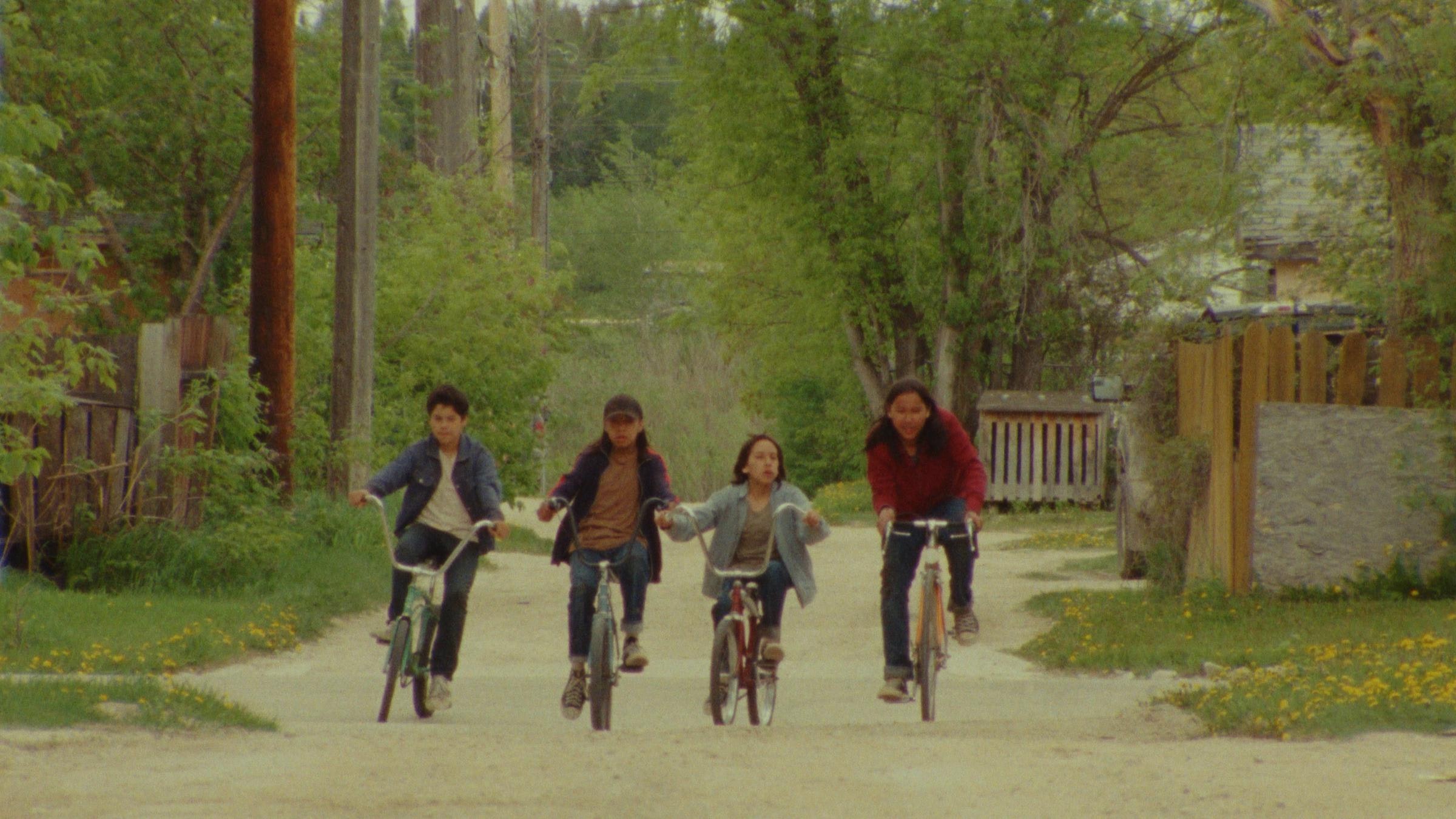 Still of four kids on bikes riding down a dirt road toward the camera. The scene is surrounded in greenery, trees, fences and telephone poles.