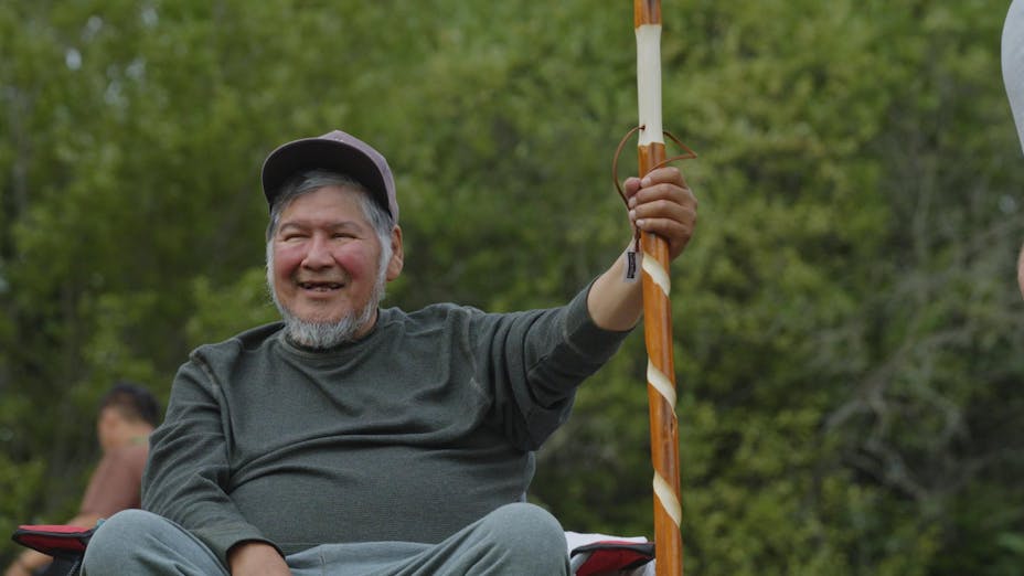 A man with a big smile and short white and gray hair and a beard wearing a hat and gray clothing sits in a camp chair and holds a carved wood staff.