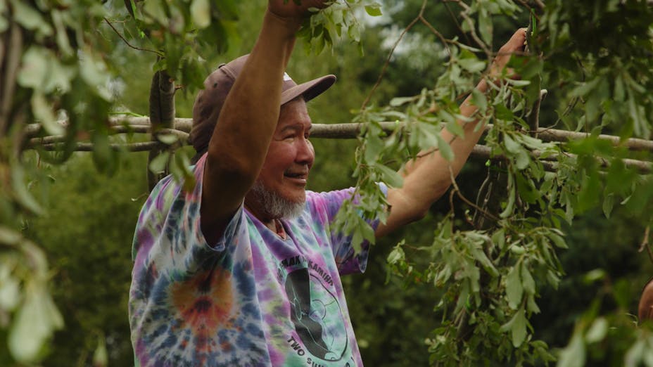 A smiling man with short white and gray hair and a beard wearing a hat and a tie dye t-shirt, with his arms up holding a structure made of branches.