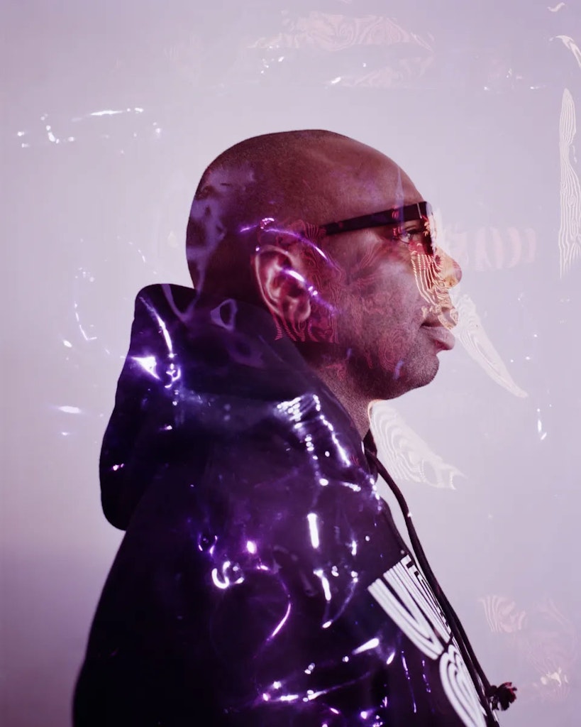 An adult in a black hoodie and glasses looks profile, face slightly obscured by light effects.