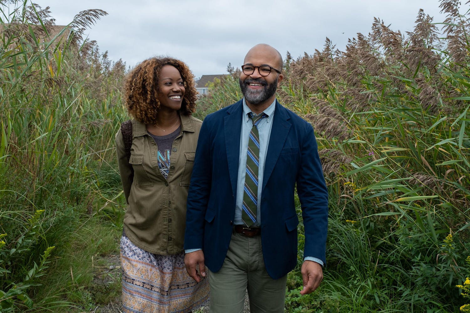 Two people, one with shoulder-length curly hair and one in a blazer and tie, smile and walk on a path in tall grasses.