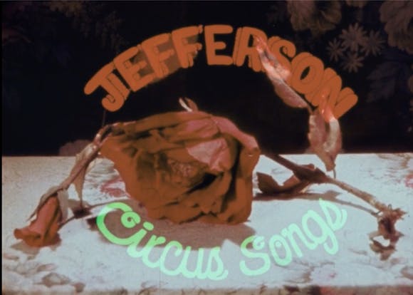 Neon sign reads "Jefferson Circus Songs" reflected in front of a red rose on a tabletop.