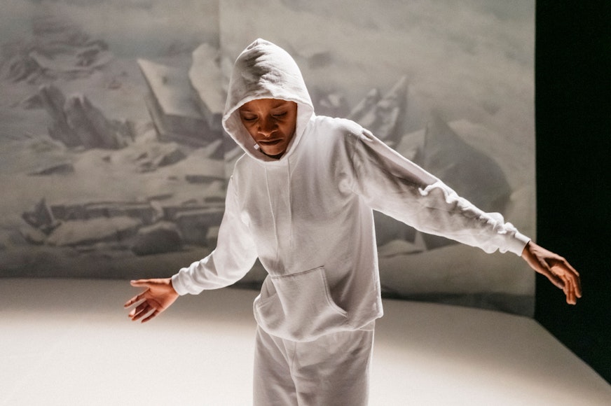 A dark-skinned man in a white-grey hoodie and pants stands against a white backdrop.