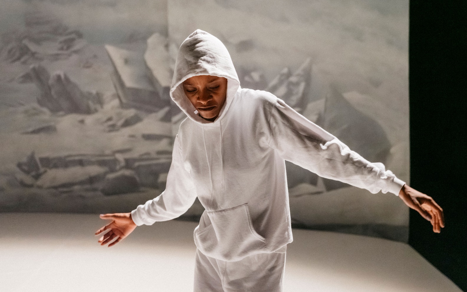 Jeremy-Nedd_From-Rock-To-Rock_Photo-Philip-Frowein_Print_10_HERO A dark-skinned man in a white-grey hoodie and pants stands against a white backdrop.