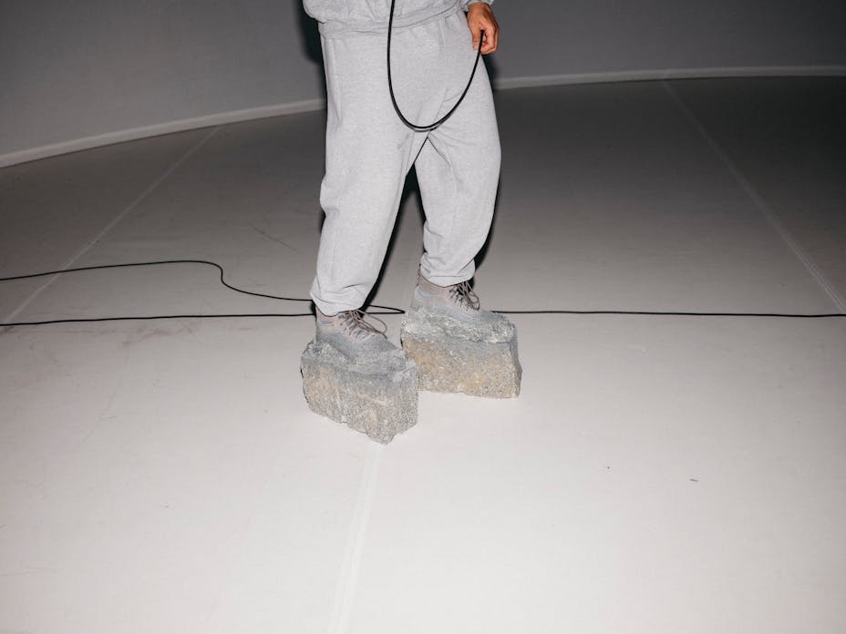 The lower half of a man with dark skin wearing grey sweatpants and shoes that are fused with giant blocks of rock.