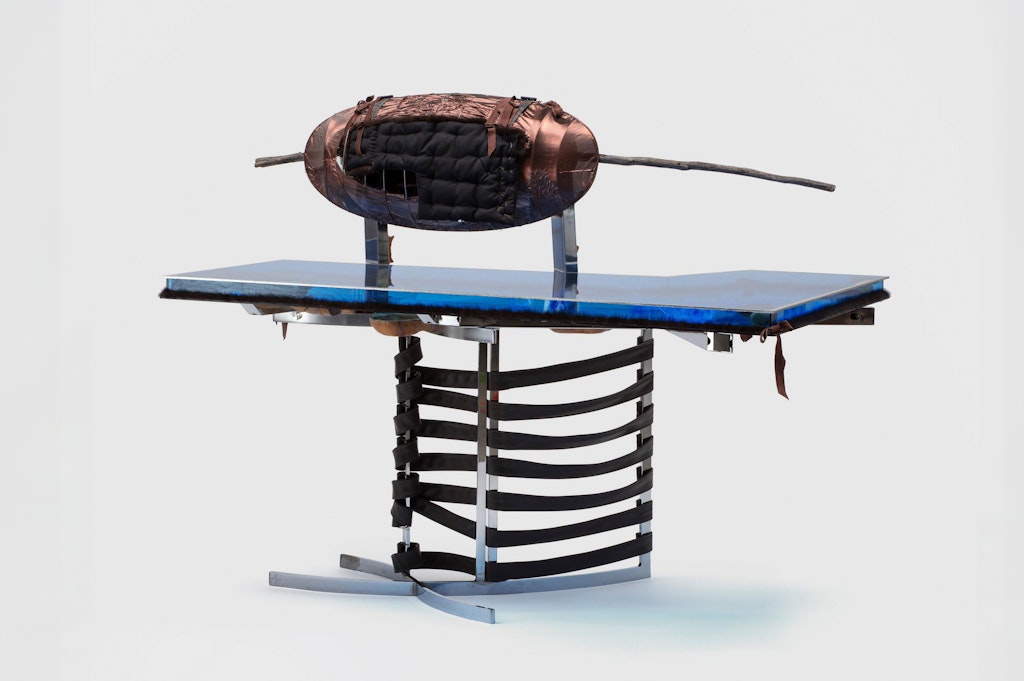 A blue acrylic tabletop resting on a metal chair with leather straps. A brown ovoid shape with a large stick behind it sits atop the table.