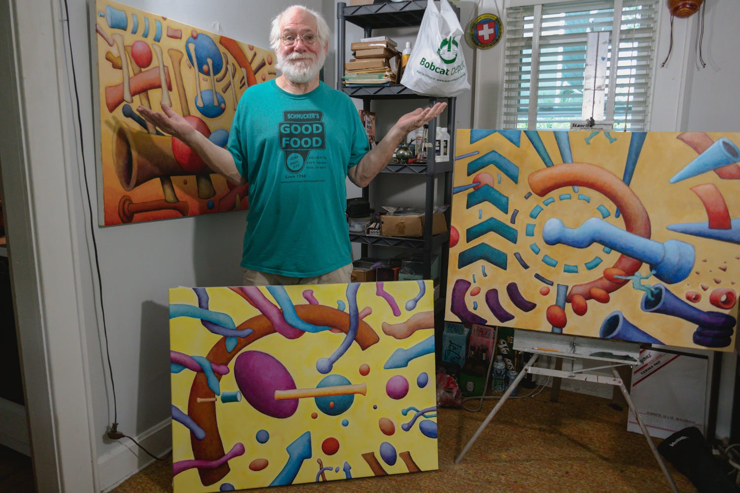 An older man with thinning white hair and beard wears round glasses and a teal t-shirt. He stands in a small room with shelves and a window behind him. There are three medium sized colorful paintings to his left, right, and in front of his legs. He holds his hands up to indicate 