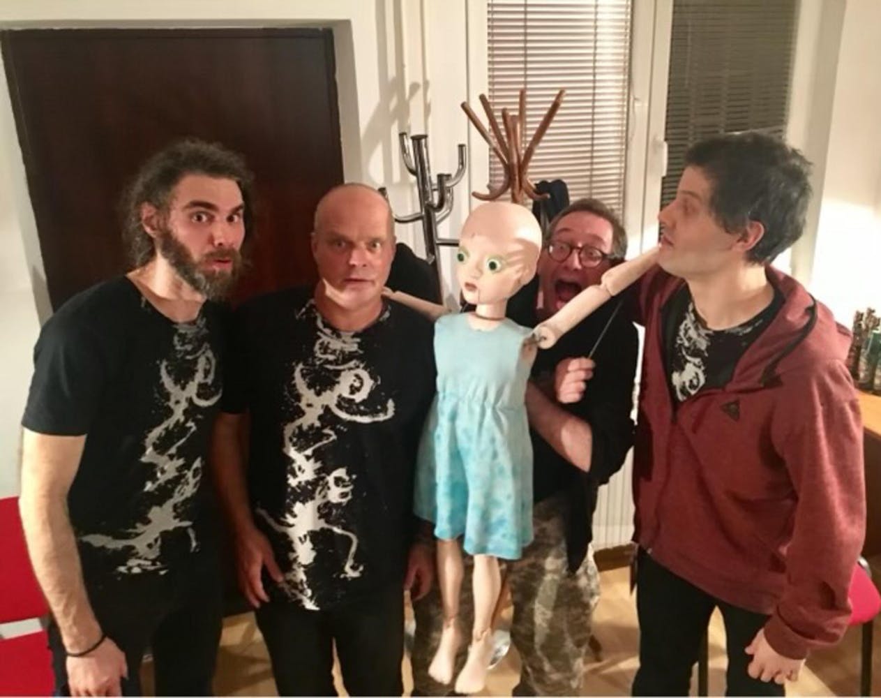 Four middle-aged men stand in a row with a large bald puppet in a blue dress in between them. Three of the men look at the camera. Three of the men wear matching black t-shirts with a white scrolling graphic. One man also wears a red jacket. One man wears a black hoodie and camo pants. They stand in an office with two windows, two coat racks, and a door behind them.