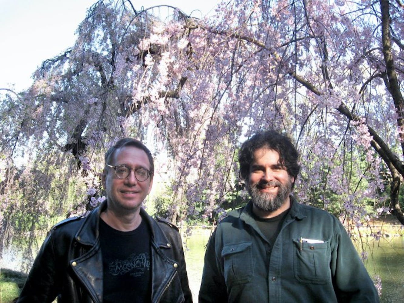 Two middle-aged men with dark hair stand in front of a large purple blossoming tree in front of a small pond. The man on the left wears glasses, a black t-shirt, and black leather jacket. The man on the right wears a graying beard, a black t-shirt, and a green overshirt. They both smile at the camera.